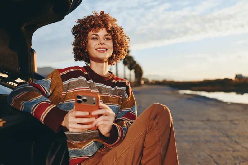 Woman with curly hair smiling in a car, holding a smartphone. Outdoor lifestyle near the seaside at sunset, warm golden light, casual travel mood, candid portrait.