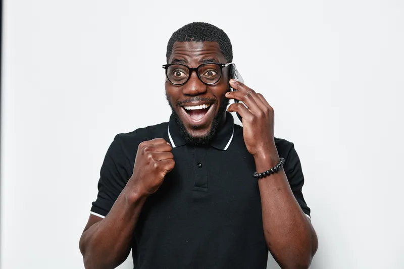 African American man with dark skin and glasses talking on phone with excited expression and fist pump gesture on white background. Casual black polo shirt and bracelet.