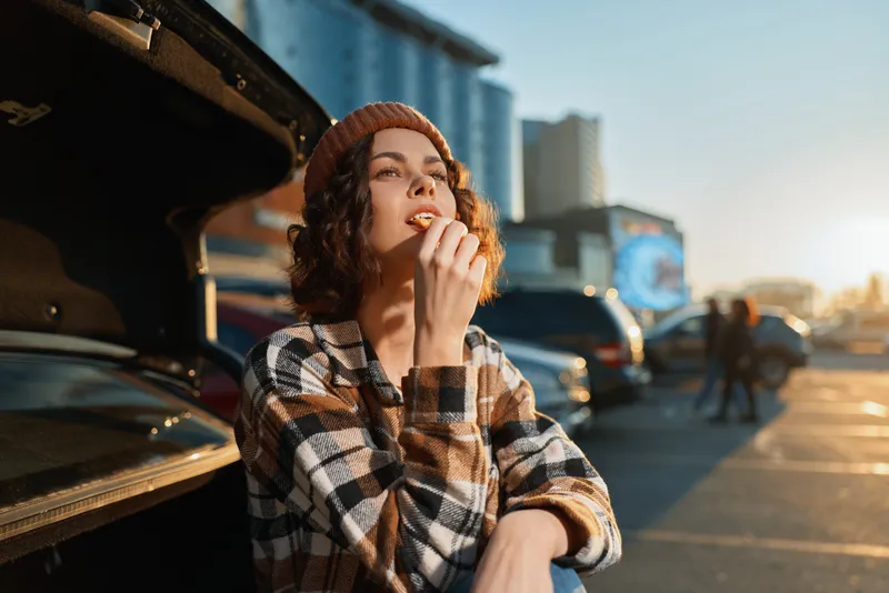 Woman car trunk snack urban beanie plaid sunset candid lifestyle portrait of a relaxed young woman eating by her open car with authenticity, mindful living and golden hour glow.