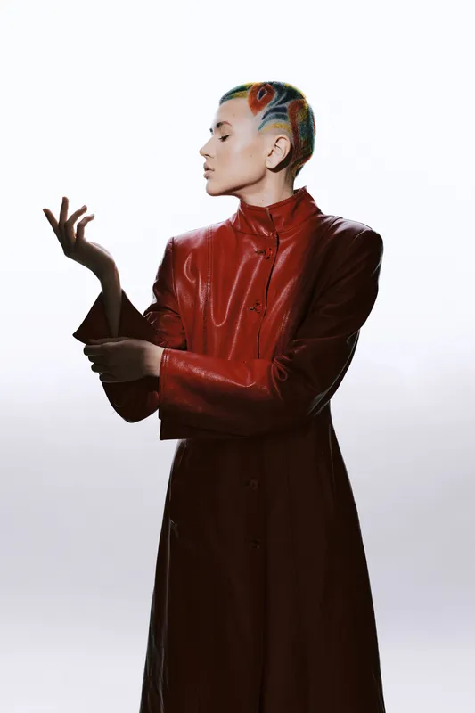 Artistic woman with colorful hair design wearing a long red leather coat, posing with elegance against a light background in a contemporary fashion style.