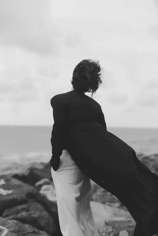 Silhouette of a figure on a rocky coastline, wind catching a flowing coat, monochrome tones, calm sea background, dramatic mood, outdoor fashion and travel vibe