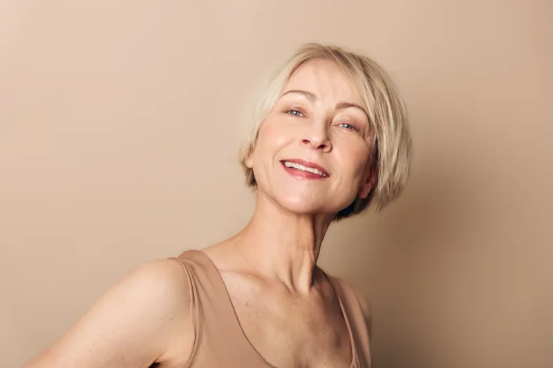 Smiling mature woman with short blonde hair wearing beige top looks confident and happy against soft beige background. Concept of aging beauty, confidence, and natural lifestyle.