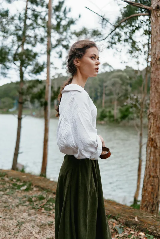 A contemplative young woman standing by the serene lake, dressed in a flowing white blouse and olive green skirt, surrounded by lush trees and nature
