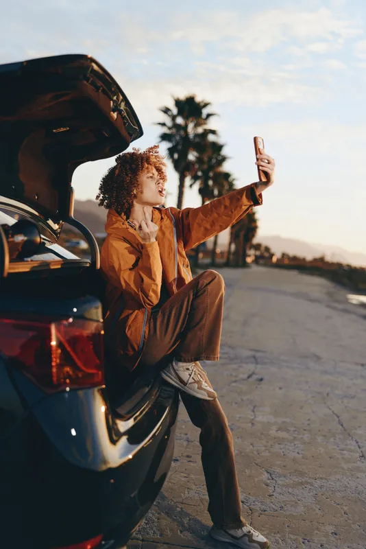 Woman lifestyle outdoors by open car trunk, taking a selfie in warm sunset light, relaxed casual pose, standing on road, palm trees softly blurred background, golden hour travel mood