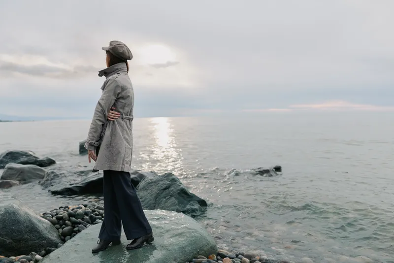 Woman in grey coat and hat stands on rocky shore looking at sea during cloudy day. Calm water and soft light create peaceful, contemplative mood by the coast.