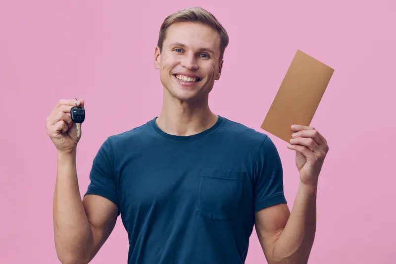 Smiling happy young man with short light brown hair wearing casual blue t-shirt holding car key and brown envelope looking at camera isolated on pink background studio portrait People lifestyle concep