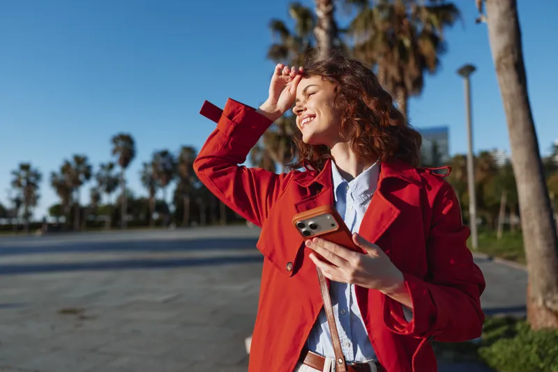 Young woman with curly hair enjoys sunny weather outdoors while holding a smartphone and smiling with her eyes closed.