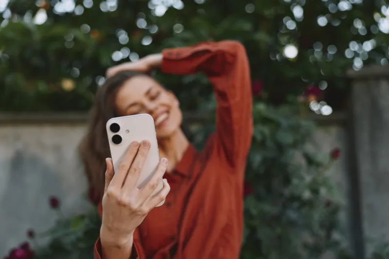 A smiling woman takes a casual selfie in a sunlit garden, wearing a rust blouse. The image captures carefree, everyday moments and confident self expression in a warm outdoor setting