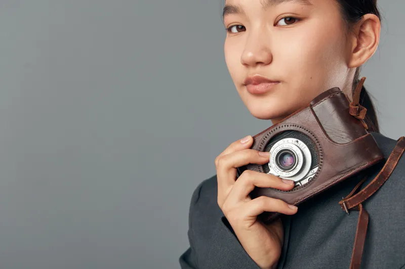 Young woman holding vintage camera close to face with leather case, portrait in studio with neutral background. Female photographer posing with retro photo equipment in casual clothing.