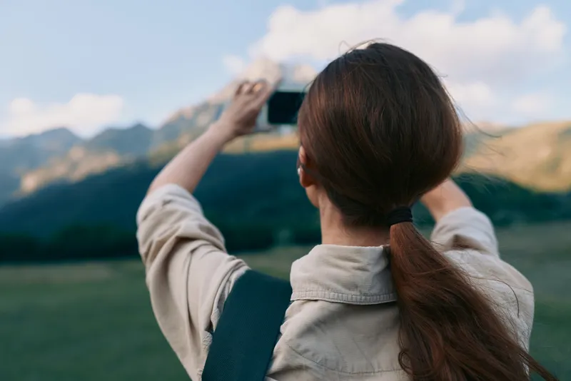 Young woman with brown hair wearing a beige jacket stands in a green field and takes a photo of distant mountains with a smartphone, creating a candid outdoor moment.