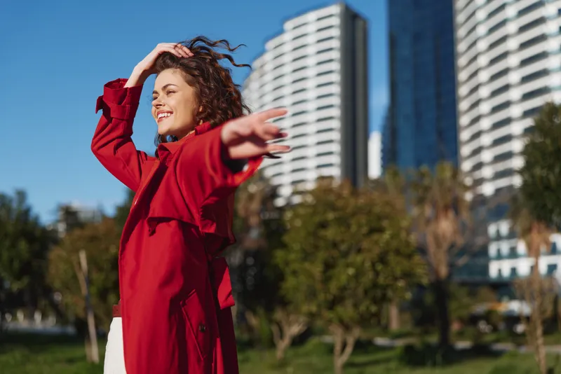 Young woman with curly hair enjoys a sunny day outdoors wearing a red coat with her arm extended and a joyful smile on her face.