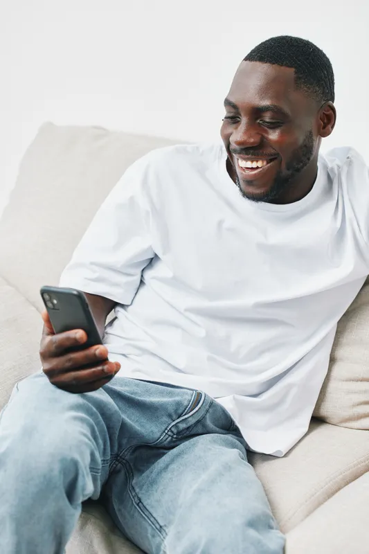 Cheerful young African man smiling while using smartphone on couch, wearing casual white t shirt and jeans, modern minimalist interior
