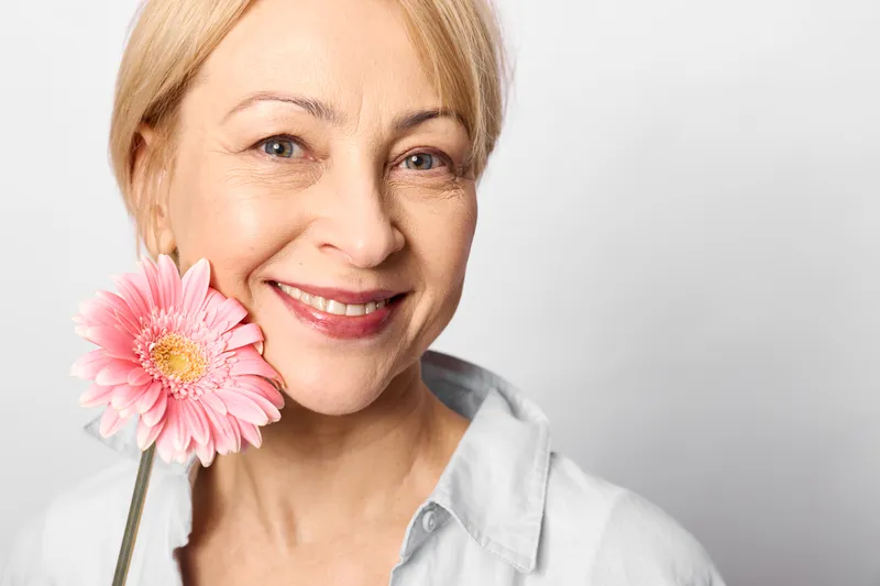 Smiling mature woman portrait, holding a pink gerbera flower near her cheek, soft studio light, neutral background, gentle beauty, spring freshness, closeup.