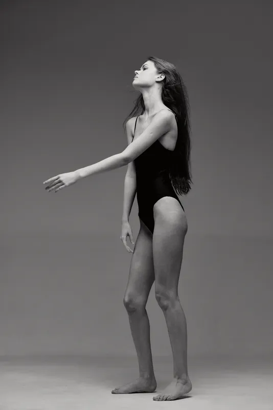 Black and white portrait of a young woman in a black swimsuit with graceful pose and long hair against a plain gray background.