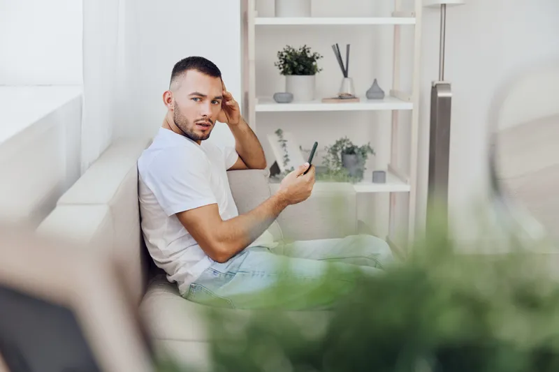 Concerned young man sitting on a couch, holding a smartphone, displaying worry. Modern interior design with plants in the background, evoking a sense of contemplation.