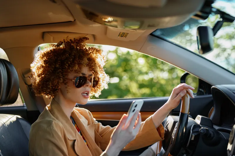 Cheerful woman driving outdoors, sunlight on curly hair, wearing sunglasses and casual blazer, checking a smartphone, retro film color mood, candid lifestyle moment, natural colors