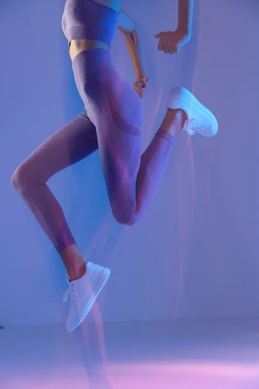 Woman sport fitness jumping in purple activewear and white sneakers with dynamic motion and cool blue lighting, capturing energy and movement in studio.