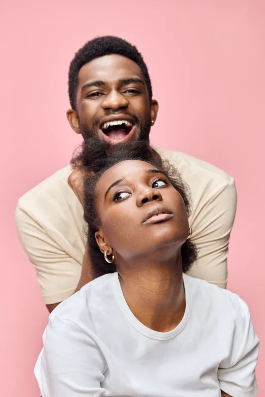 Joyful young couple with playful expressions against a soft pink background, showcasing love, connection, and happiness in their casual outfits