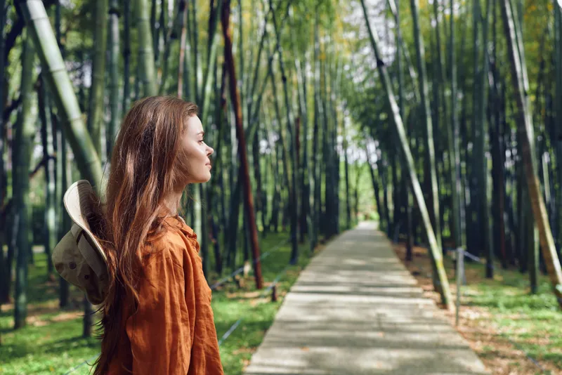 Woman, bamboo, path, forest, hat, profile, nature, travel. Young woman in profile wearing a straw hat and rust jacket walking along a paved path through a dense bamboo forest, peaceful journey.