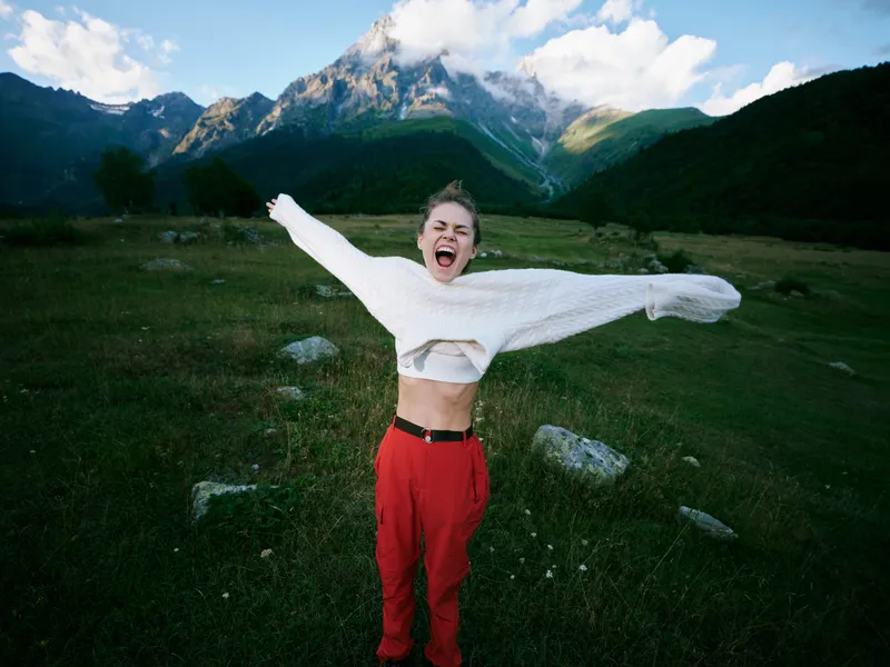 Outdoor celebration in alpine landscape, a joyful figure spans arms wide, wearing a white sweater and red pants, with mountains and green valley in the background