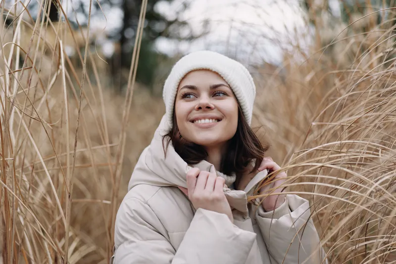 Winter outdoors portrait of a smiling woman in a light coat and beanie, standing among tall grasses in a field, conveying warmth, serenity and beauty.