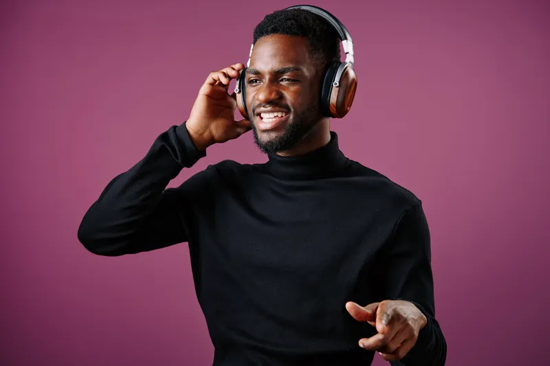 Young Black man joyfully listening to music with headphones, vibrant purple background showcasing trendy fashion and positive emotions