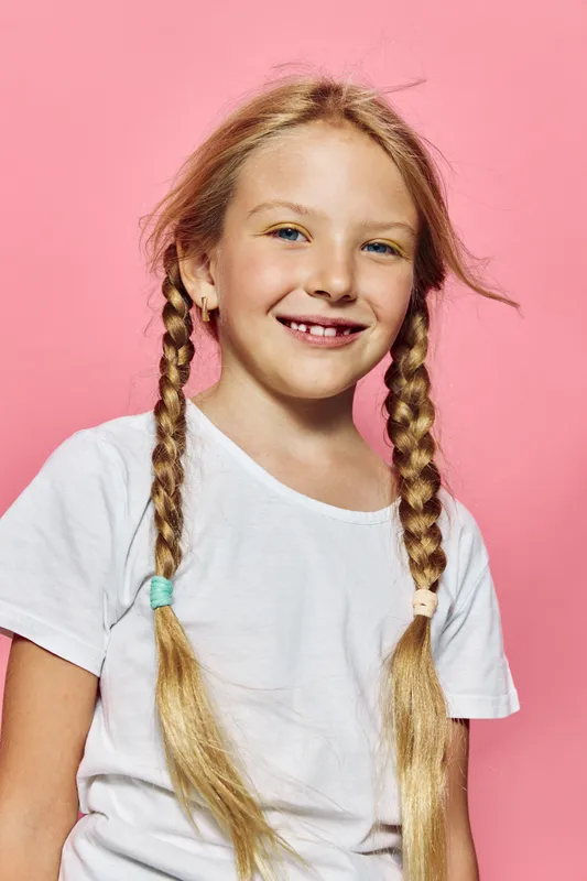 Smiling young girl with blonde braided hair and missing front teeth wearing a white t-shirt posing against a pink background. Portrait of happy childhood and innocence.