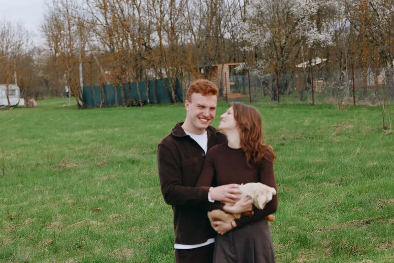 Smiling couple in a field gently holds a goat kid, enjoying a casual outdoor moment. Warm natural light and relaxed mood highlight companionship, animals, and everyday happiness in a countryside