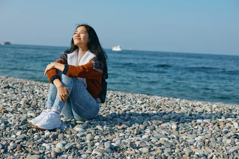 A serene beach scene with a woman seated on pebbles by the sea, wearing a cozy sweater and denim, gazing toward the horizon in a quiet coastal mood.