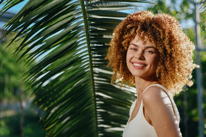 Smiling woman with curly hair enjoys sunny tropical outdoors near palm leaves, radiating happiness, natural beauty, and warm summer vibes in casual white tank top.