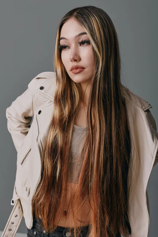 Young woman with long straight hair wearing light jacket and tank top posing with hands on hips against gray background for fashion and beauty portrait.