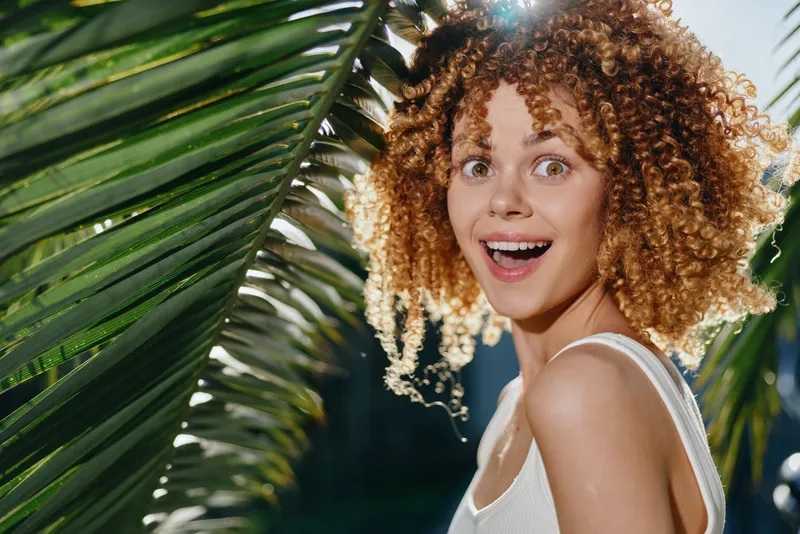 happy woman with curly hair smiles brightly outdoors near tropical palm leaves in sunny natural light wearing white tank top, capturing joyful summer vibe