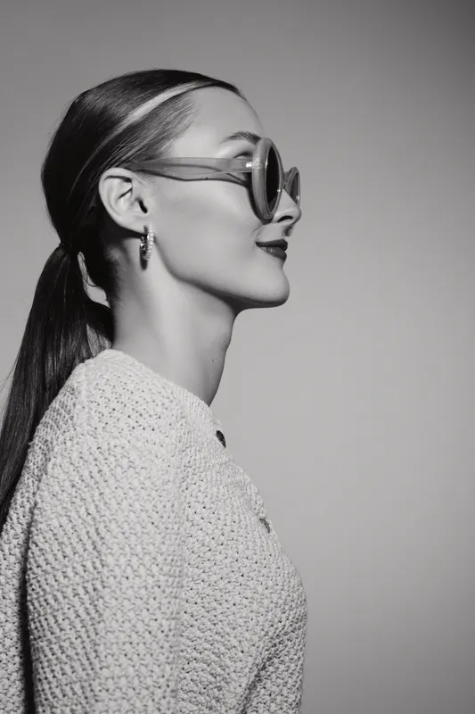 Profile of a Caucasian woman with pale skin wearing sunglasses and knitted sweater on grey background.