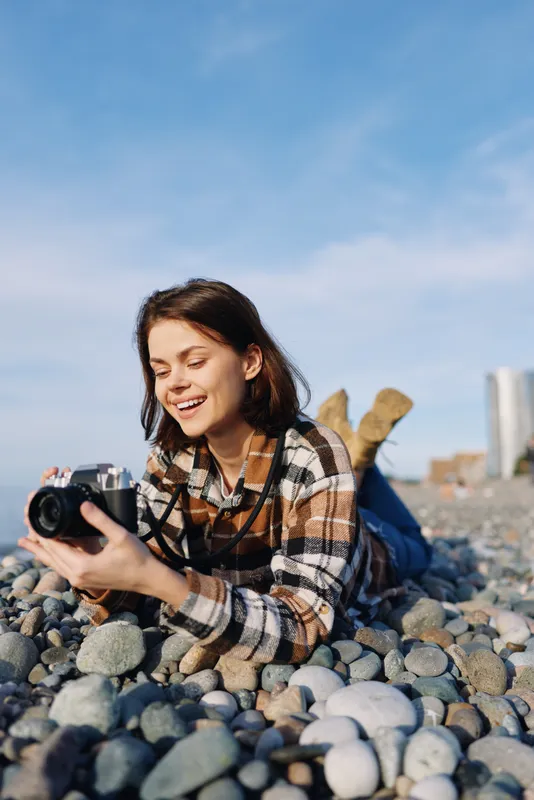 Woman on a rocky beach with camera, smiling and focused on photography, wearing a plaid shirt. Casual outdoor scene by pebbles and blue sky, capturing a relaxed coastal moment.