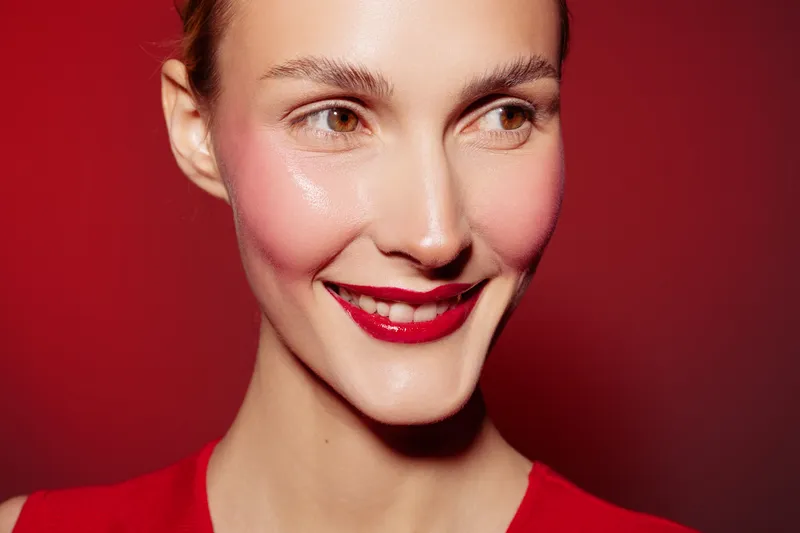 Close-up of smiling woman with red lipstick and glowing skin on red background. Concept of beauty, makeup, skincare, and confident female portrait with vibrant colors and studio lighting.