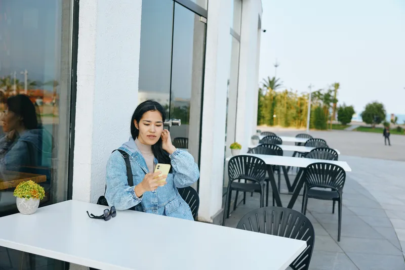 Woman sits at an outdoor cafe using a smartphone beside sunglasses on a white table, blue jacket, bright day, urban setting, relaxed moment ideal for communication and leisure
