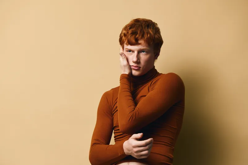 Young man with red hair and freckled skin wearing brown turtleneck posing thoughtfully against beige background with hand on face and calm expression.
