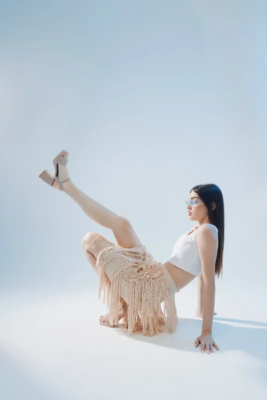 Young woman in stylish summer outfit poses with one leg raised on white background. Concept of beauty, fashion, and confident female model in studio lighting.