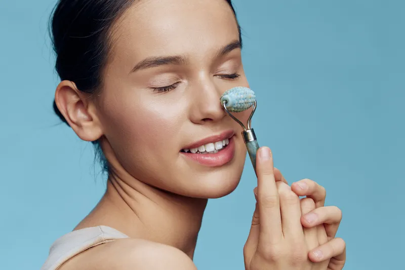 Young woman using a facial roller on her cheek, promoting relaxation and skincare The soothing blue background enhances the serene atmosphere