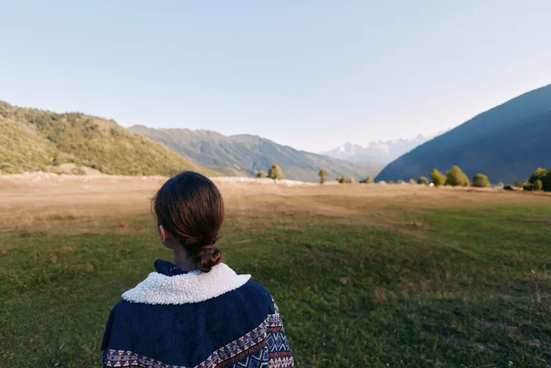 person woman mountains valley meadow nature back landscape in this photo: a woman seen from behind stands in a wide grassy meadow looking toward distant mountain valley under clear sky