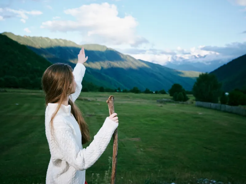 Outdoor scene with a girl in a white sweater raising a wooden stick toward distant mountains across a green valley, blue sky and peaceful countryside, capturing exploration, nature and adventure