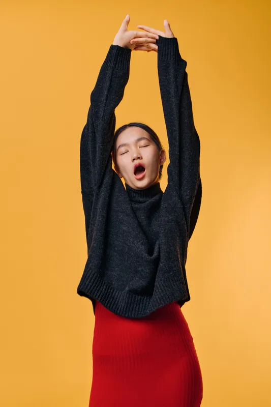 Young woman yawning with eyes closed stretching arms up in cozy black sweater and red skirt on orange background. Relaxed tired female model expressing fatigue and comfort at studio.