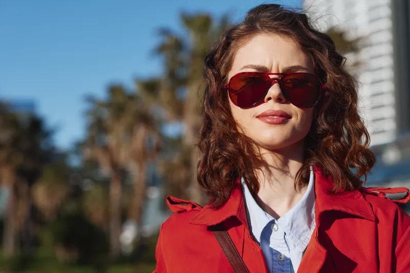 A stylish woman with short wavy hair wears a red trench and brown bag, posing confidently outdoors with palm trees and city buildings in the background on a sunny day.