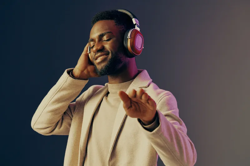 Young African man enjoying music with headphones, wearing a stylish light coat, immersed in sound against a gradient background, capturing joy and relaxation