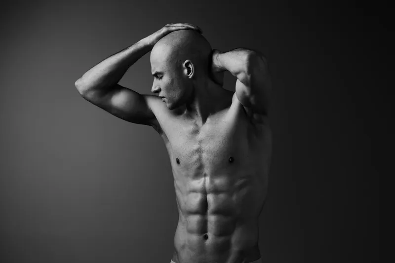 Black and white image of a muscular man posing confidently, showcasing his toned physique and strong silhouette against a dark background.