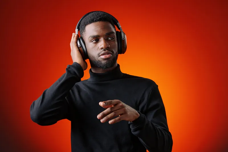 Young African American man enjoying music with headphones, wearing a stylish black turtleneck against a vibrant orange background