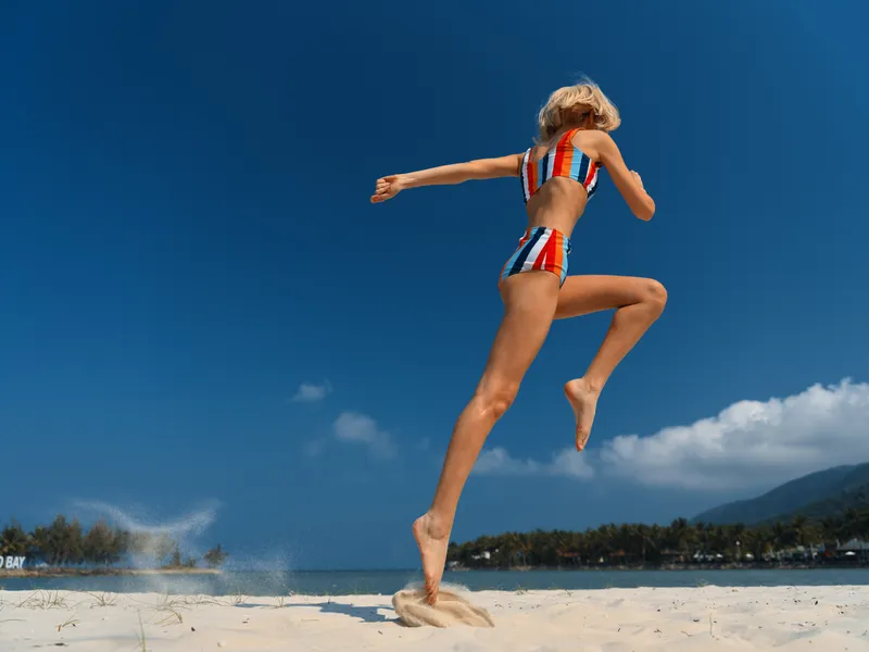 Dynamic action shot of a person mid jump on a sunny beach, wearing a striped bikini, capturing motion, energy, and carefree summer fun against a crisp blue sky.