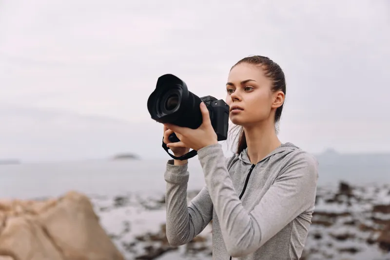 Young woman photographing coastline with camera, wearing gray athletic outfit, showcasing focus and determination near the sea