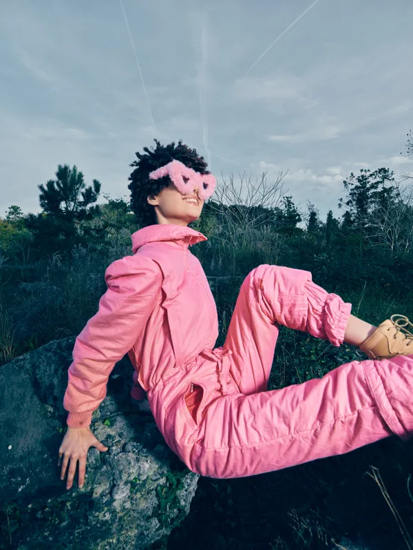 Pink jumpsuit worn by a person posing outdoors on rocks, wearing heart-shaped sunglasses, capturing a bold fashion moment in natural daylight.