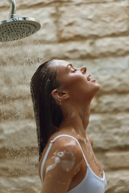 woman showering outdoors with water cascading, enjoying refreshing moment, natural tan skin, wet hair, white tank top, textured stone wall background